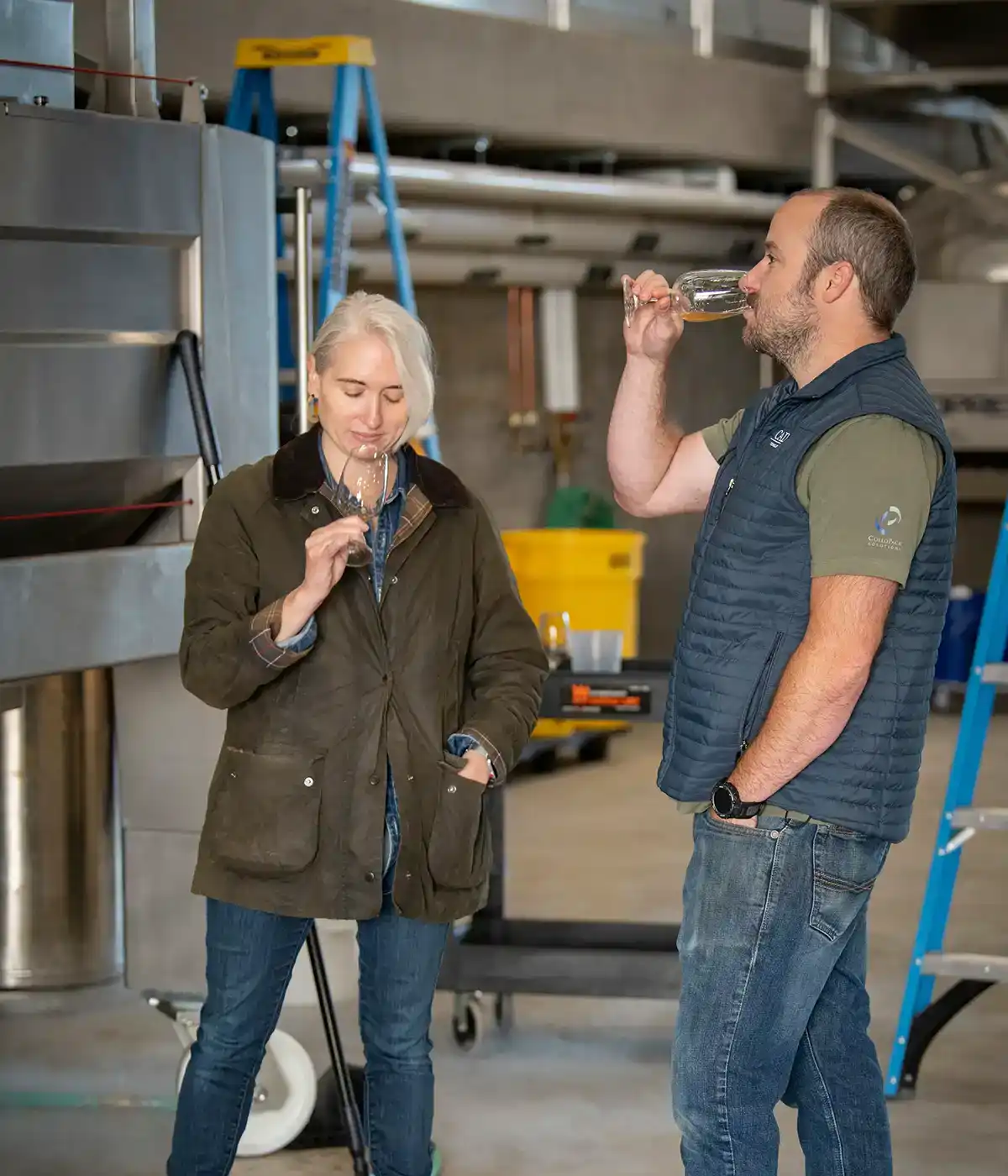 Two people in casual attire tasting Chardonnay wine in an industrial setting. One is holding a glass to their nose while the other takes a sip, savoring the flavors of Willamette Valley. They stand next to large stainless equipment and a yellow bin.