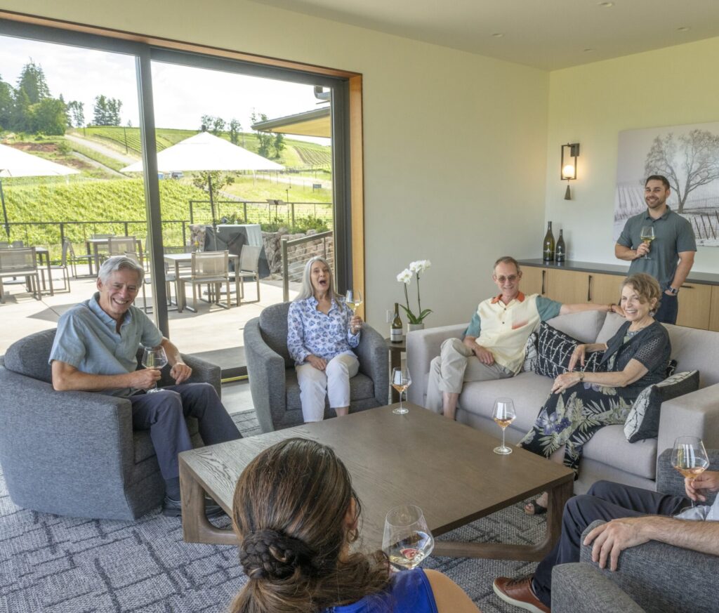 A group of adults sitting in a modern living room, holding glasses and engaging in conversation. A patio and green landscape are visible through large glass doors in the background.