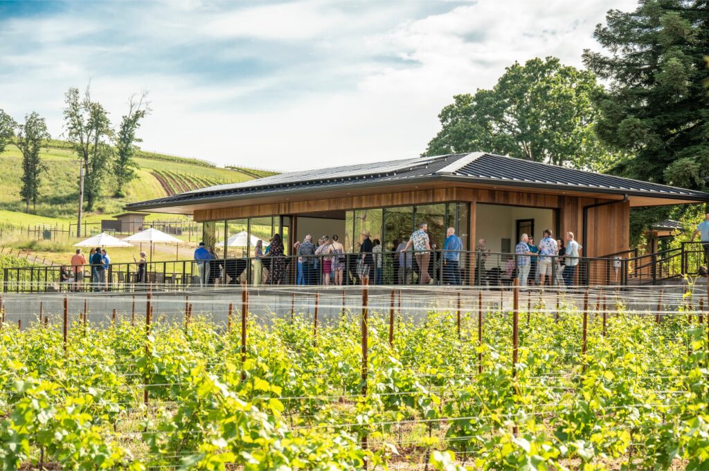 A group of people gather on a modern building's deck overlooking a lush vineyard under a partly cloudy sky.